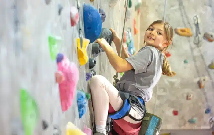 Child using a harness while climbing a colorful indoor rock wall, showcasing climbing wall options for schools and recreation centers of different sizes.